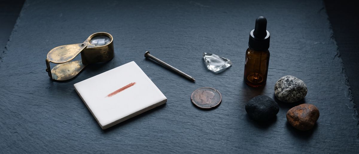 The guides' kit laid out on slate: a loupe, a porcelain tile with a red streak, a nail, a copper coin, a shard of glass, a dropper bottle and three pebbles.